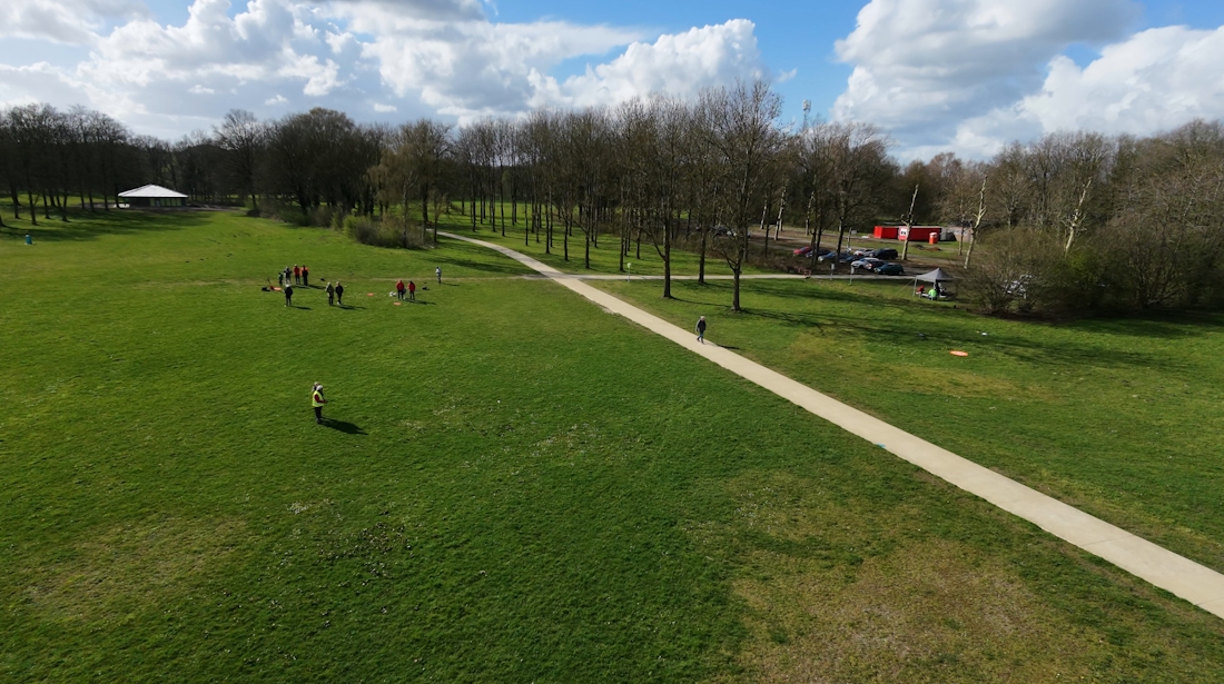 Een luchtfoto van een groot groen grasveld in een park op een zonnige dag met wolken in de lucht. Over het veld loopt een licht gekleurd wandelpad dat schuin door het beeld loopt. Verspreid over het gras staan kleine groepjes mensen, waarschijnlijk bezig met een activiteit of sport. Aan de rechterkant van de foto staan bomen zonder bladeren en daarachter is een parkeerplaats met auto’s te zien en een rood gebouw. Links op de achtergrond staat een open paviljoen. Het geheel geeft een rustige, open parkachtige omgeving weer gezien vanuit de lucht, waarschijnlijk gemaakt met een drone.