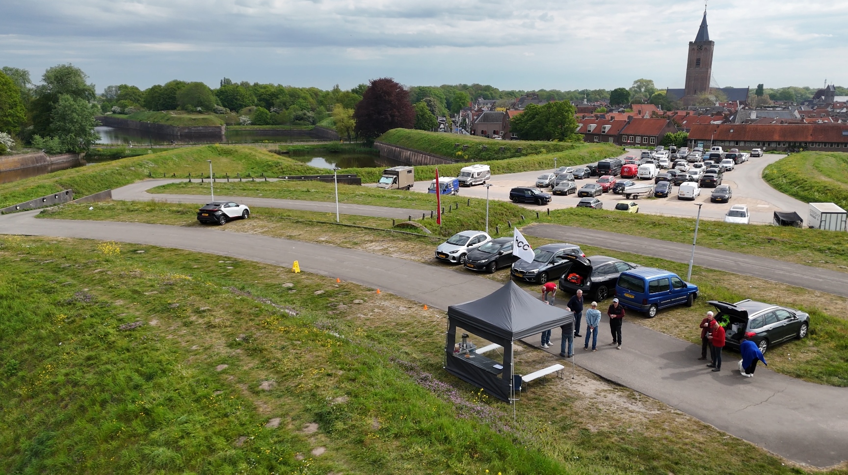 Luchtfoto van een dronebijeenkomst bij de vesting van Naarden. Op de voorgrond staat een kleine partytent langs een weg, waar enkele mensen bij elkaar staan en praten. Naast de tent staan meerdere geparkeerde auto’s met open achterkleppen. In de omgeving is veel groen gras en een vestingwal met water eromheen. Op de achtergrond is een druk parkeerterrein te zien en daarachter de historische gebouwen van Naarden, met een grote kerk die boven alles uitsteekt. Het weer is licht bewolkt met af en toe zon.